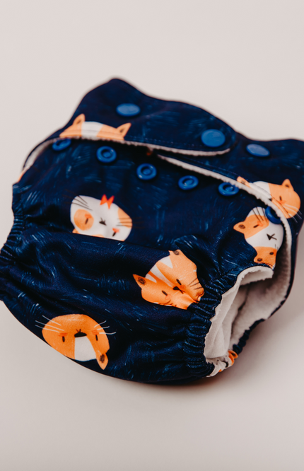 Reusable cloth diaper showcasing side snaps and soft elastic leg openings for a snug fit. Adorable cat print on navy blue fabric. breathable material for comfort and safety. Perfect for eco-conscious parents looking for a sustainable, stylish diaper.