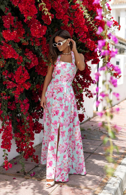 Model wearing a multicolor floral maxi dress with wide neck-tie straps and side slit, standing near vibrant red bougainvillea. Summer-ready LaBalancia daydress for warm weather and vacations. Elegant sleeveless design, perfect for stylish getaways and garden parties.